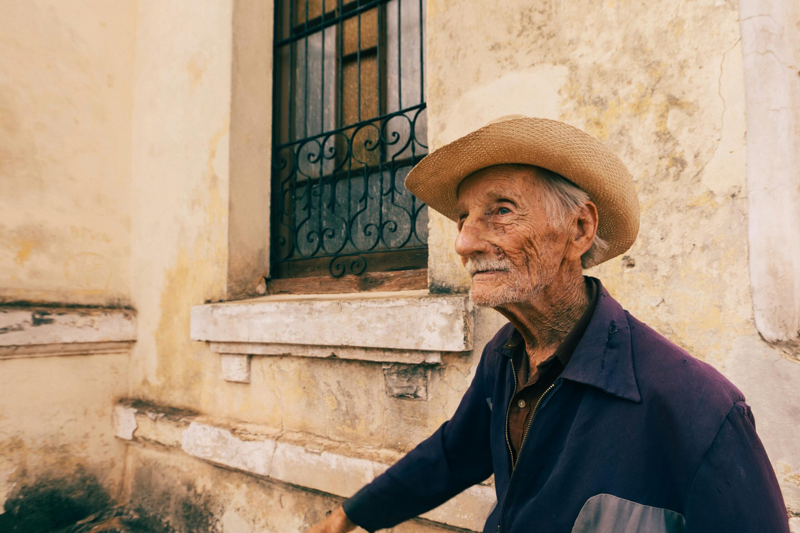 Elderly man wearing a straw hat stands by a weathered wall, showcasing character and history.