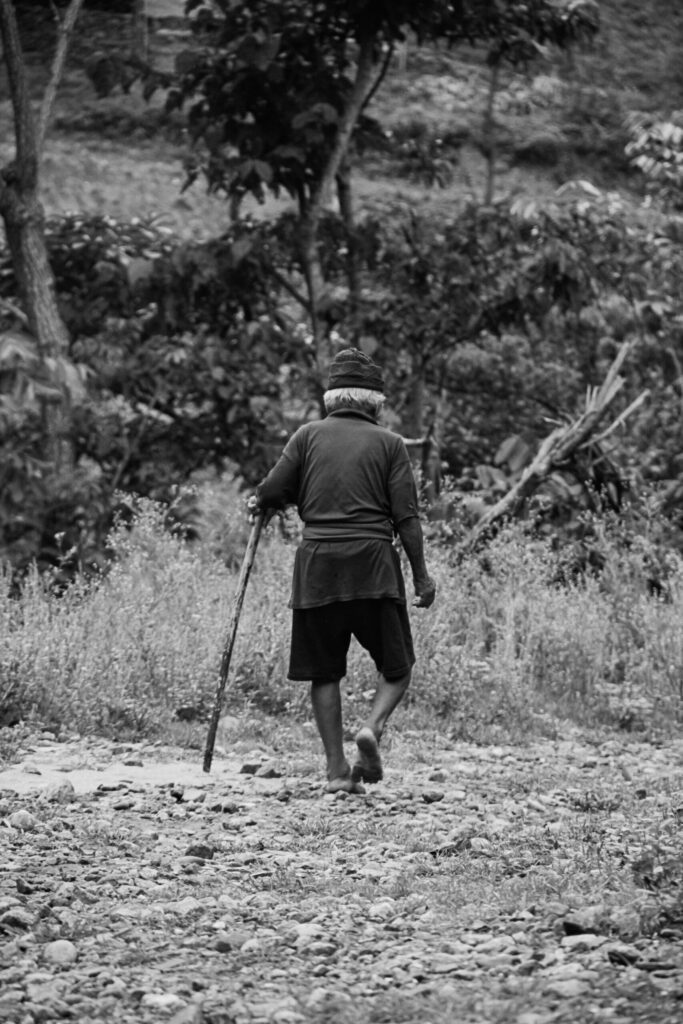Monochrome photo of an elderly man walking with a stick along a rural path in Nepal.